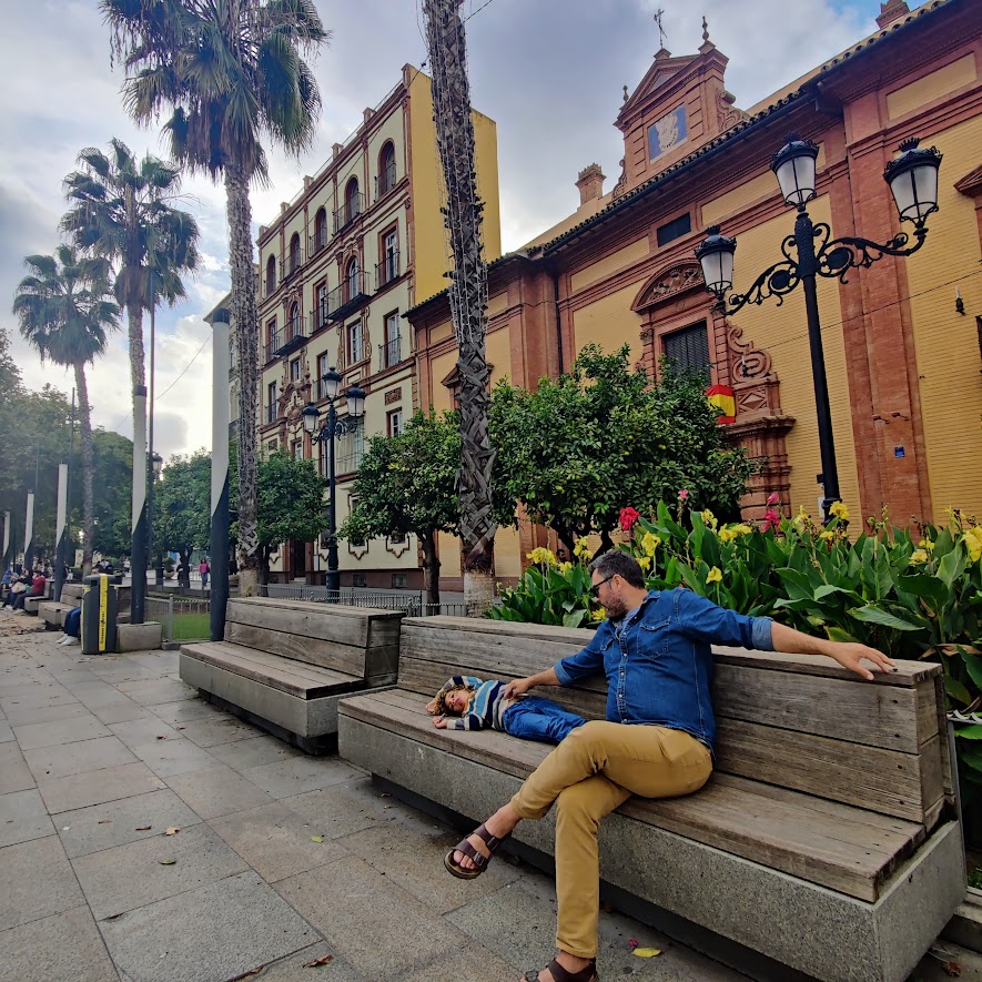 picture of a man sitting on a bench, with a child laying down on the bench, with palm trees and Spanish style buildings in the back